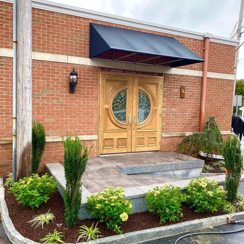 Entrance of a brick building with wooden double doors, a black awning, and landscaped greenery in front.