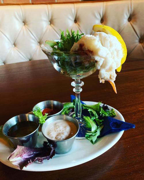 A glass dish with shrimp cocktail, topped with lettuce and a lemon wedge, served with three dipping sauces on a plate.
