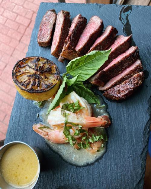 Sliced steak on a slate plate, accompanied by grilled lemon, shrimp, basil, and a small cup of sauce.