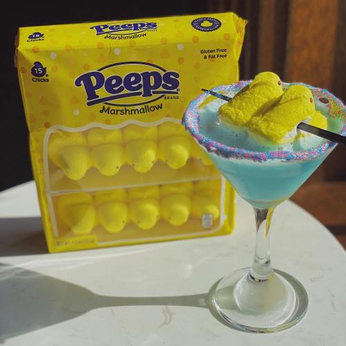 A colorful cocktail topped with yellow marshmallow chicks, next to a box of Peeps marshmallows on a sunny surface.