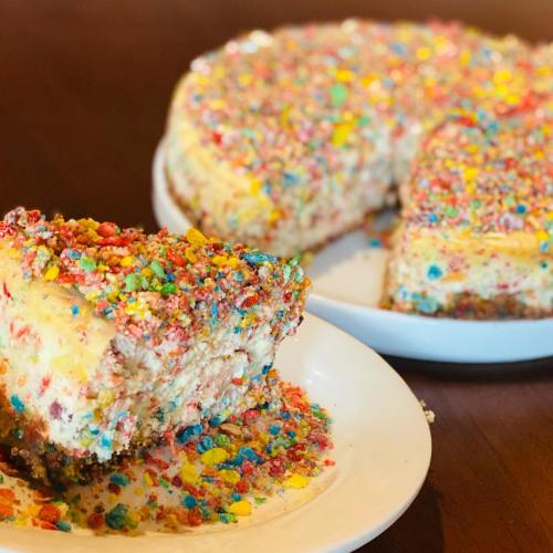 A slice of colorful cake topped with sprinkles, sitting on a white plate with the rest of the cake in the background.