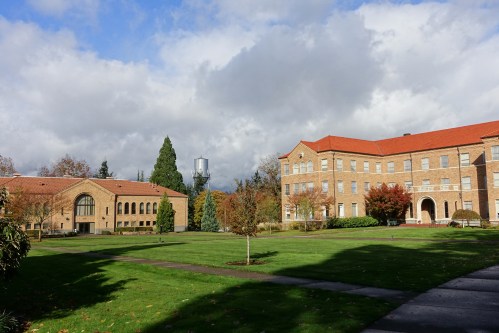 A campus scene featuring brick buildings, green lawns, and a cloudy sky. Trees and a water tower are visible in the background.