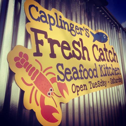 Sign for Caplinger's Fresh Catch Seafood Kitchen, featuring a lobster and fish graphic, open Tuesday to Saturday.