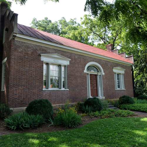 A brick building with a red roof, white windows, and a wooden door, surrounded by greenery and flowers.