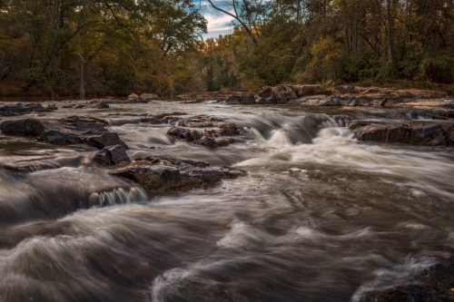 A serene river flows over rocky terrain, surrounded by trees in autumn colors under a cloudy sky.