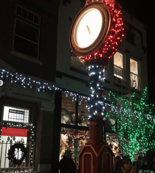 A decorative clock adorned with colorful lights stands outside a shop, surrounded by festive holiday decorations.