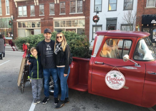 A family poses by a vintage red truck loaded with a Christmas tree in a festive town square.