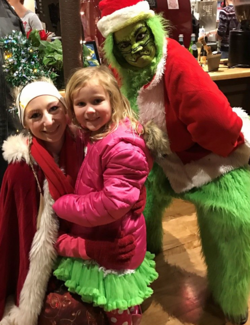 A girl in a pink coat poses with a woman in festive attire and a person dressed as the Grinch.