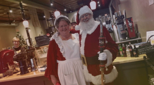 A woman in a white dress and apron stands next to a man in a Santa costume at a festive café.