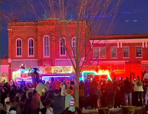 Crowd gathered at night watching a festive parade with colorful lights and a decorated fire truck.