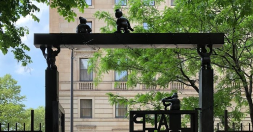 Sculpture of three figures atop a black archway, surrounded by green trees and a building in the background.