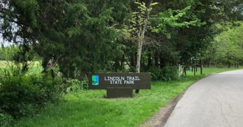 Sign for Lincoln Trail State Park beside a winding road, surrounded by green trees and grass.