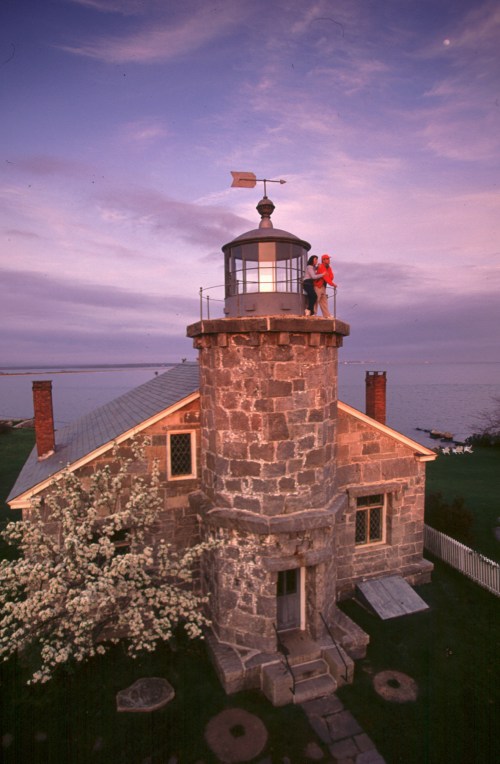A stone lighthouse at sunset, with two people on top and blooming trees nearby, overlooking a calm body of water.