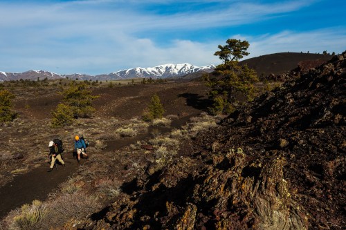 Two hikers walk along a trail in a volcanic landscape, with snow-capped mountains in the background and sparse vegetation.