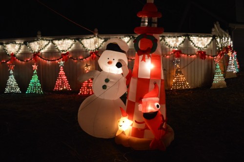 A festive scene featuring a snowman and a lighthouse, surrounded by colorful Christmas lights and decorated trees.