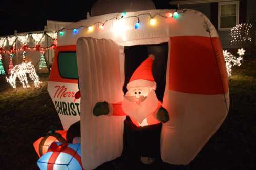 A festive inflatable Santa stands in a decorated doorway, surrounded by colorful Christmas lights and gifts.