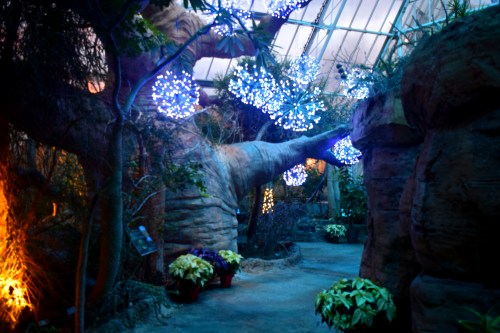A serene indoor garden with glowing blue lights, large trees, and potted plants lining a winding path.