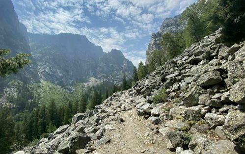 A rocky trail winds through a mountainous landscape, surrounded by trees and steep cliffs under a cloudy sky.