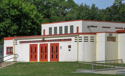 Exterior of a swimming pool building with red doors and a white facade, surrounded by green grass and trees.
