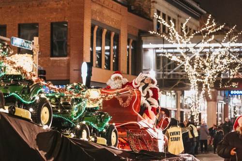 Santa and Mrs. Claus wave from a decorated float during a festive parade, surrounded by twinkling lights.