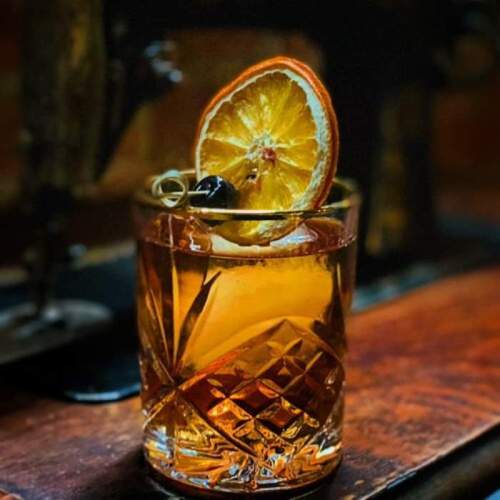 A crystal glass filled with a brown cocktail, garnished with a dried orange slice and a cherry.