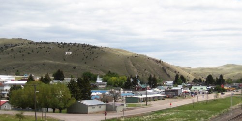 A small town nestled in a valley, surrounded by green hills and dotted with trees under a cloudy sky.