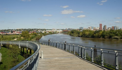 A scenic view of a river with a wooden walkway, surrounded by greenery and a city skyline in the background.