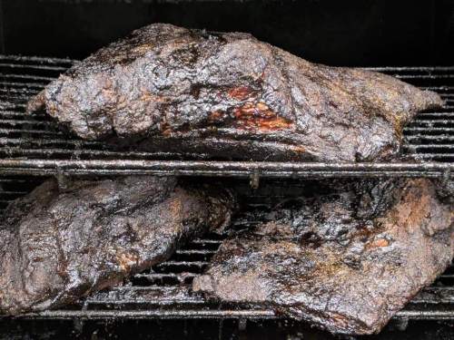Three large, smoked briskets resting on grill racks, showcasing a dark, caramelized exterior.
