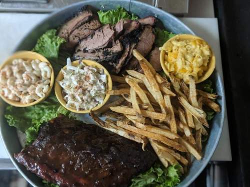 A platter featuring smoked meats, coleslaw, macaroni and cheese, shrimp, and crispy fries on a bed of lettuce.