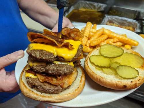 A towering burger with multiple beef patties, cheese, bacon, and pickles, served with crinkle-cut fries on a plate.