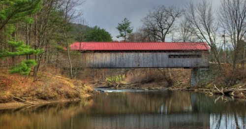 A wooden covered bridge with a red roof spans a calm river, surrounded by trees and a cloudy sky.