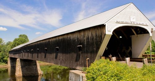 A historic covered bridge with a wooden structure, spanning a calm river surrounded by greenery and blue skies.
