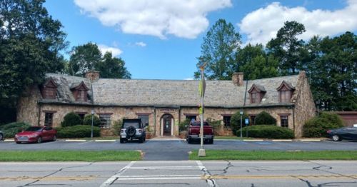 A stone building with a sloped roof, surrounded by trees, and several parked cars in front on a sunny day.