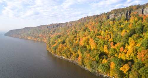 A scenic view of a riverbank lined with vibrant autumn foliage and rocky cliffs under a partly cloudy sky.