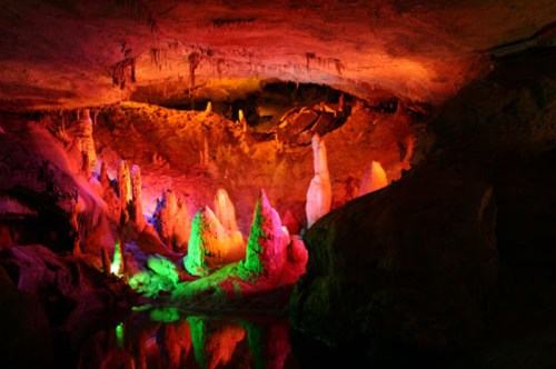 Colorful illuminated stalagmites and stalactites in a cave, reflecting in still water below.