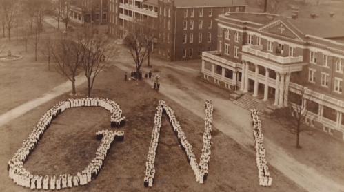 Aerial view of a large group of people forming the letters "GNI" on a grassy area near historic buildings.