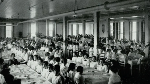 A historic dining hall filled with rows of women in white uniforms seated at long tables.