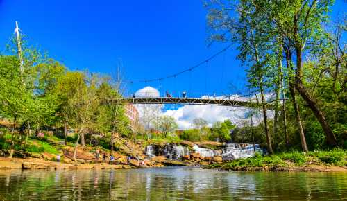 A scenic view of a river with a waterfall, surrounded by greenery and a pedestrian bridge overhead under a blue sky.