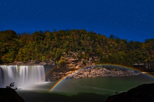 A serene waterfall under a starry night sky, with a vibrant rainbow arching over the calm water.