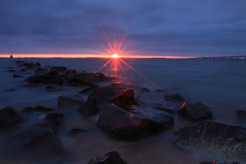Sunset over the ocean, with rays shining through clouds and reflecting on water, surrounded by rocky shoreline.