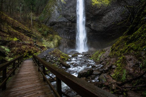 A wooden bridge leads to a waterfall cascading down rocky cliffs, surrounded by lush greenery and mist.