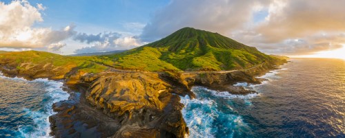 A panoramic view of a lush green mountain by the ocean, with rocky shores and waves crashing against the coastline.