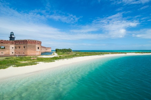 A scenic view of a historic fort by the beach, with clear turquoise water and a blue sky in the background.
