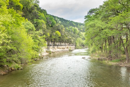 A serene river flows through lush green trees and rocky cliffs under a cloudy sky.