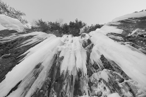 Icicles hang from rocky cliffs, surrounded by snow and sparse vegetation, under a clear sky. Black and white image.
