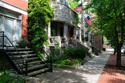 A tree-lined street featuring stone buildings with steps, planters, and American flags hanging from the balconies.