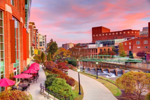 A scenic view of a riverwalk at sunset, featuring colorful buildings, greenery, and outdoor seating with pink umbrellas.