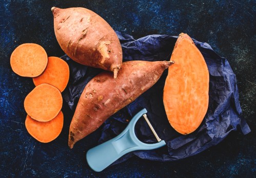 A flat lay of sweet potatoes, some whole and sliced, with a blue peeler on crumpled dark paper.