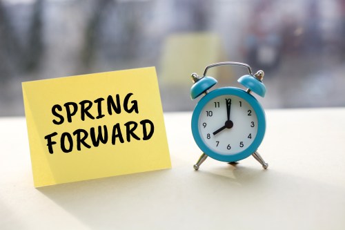 A blue alarm clock next to a yellow sticky note that says "SPRING FORWARD."
