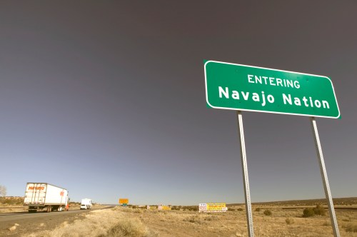 A green road sign reads "Entering Navajo Nation" with trucks on the highway and a clear sky in the background.